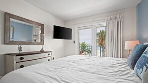 A bedroom with a large bed, a dresser, a flat-screen TV, a balcony with curtains, and a view of palm trees. - Direct Gulf Front 2BR/2BA condo (Gulf Shores)