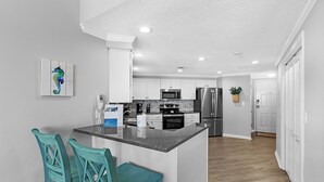A modern kitchen with a central island, white cabinetry, stainless steel appliances, and teal chairs. - The Palms 501 by Liquid Life (Orange Beach)
