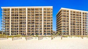 A beachfront hotel with multiple balconies, a sandy beach in front, and a clear blue sky. - The Palms 501 by Liquid Life (Orange Beach)