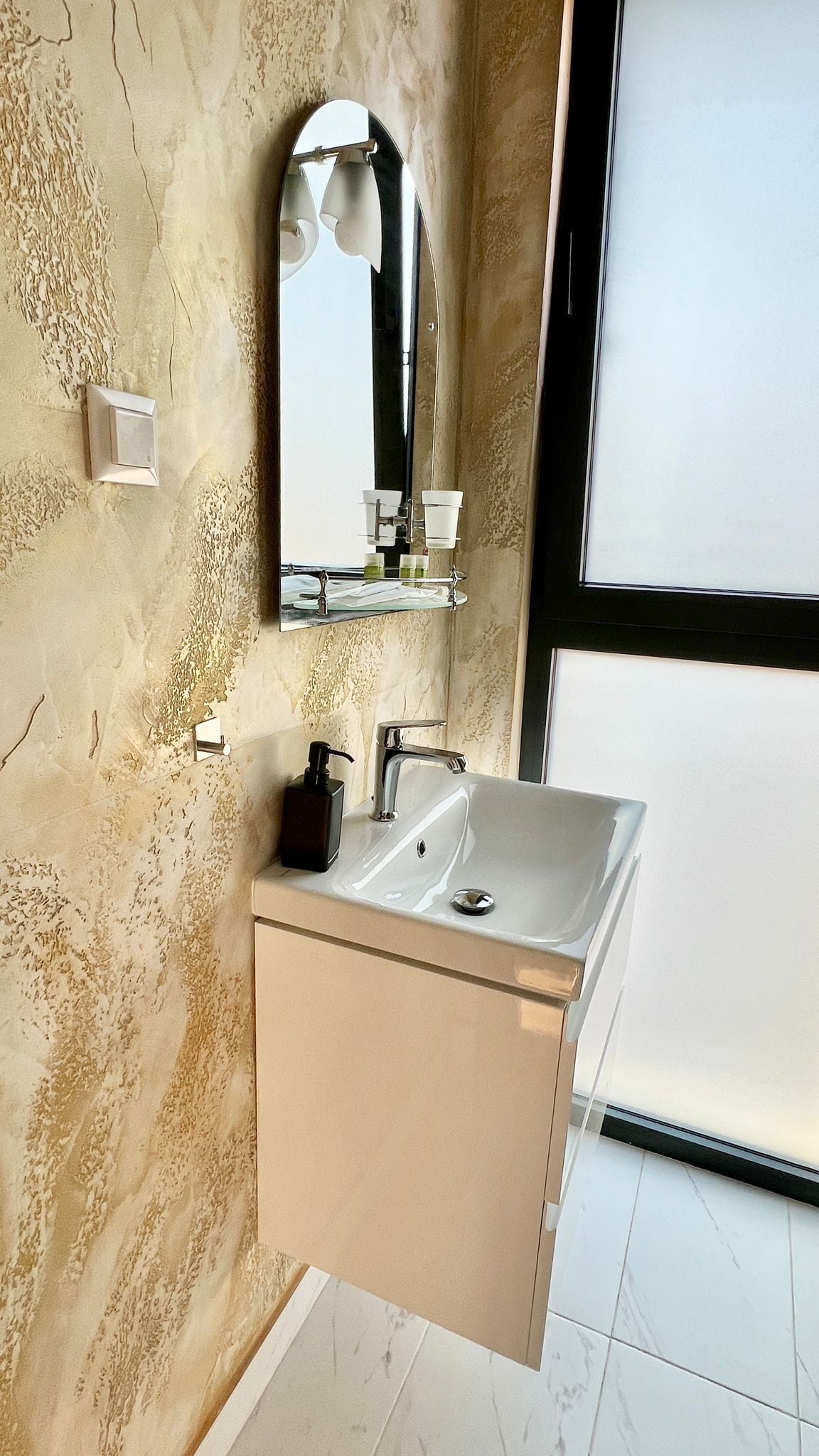 Un baño con pared de mármol, un espejo grande, un lavamanos y un dispensador de jabón.