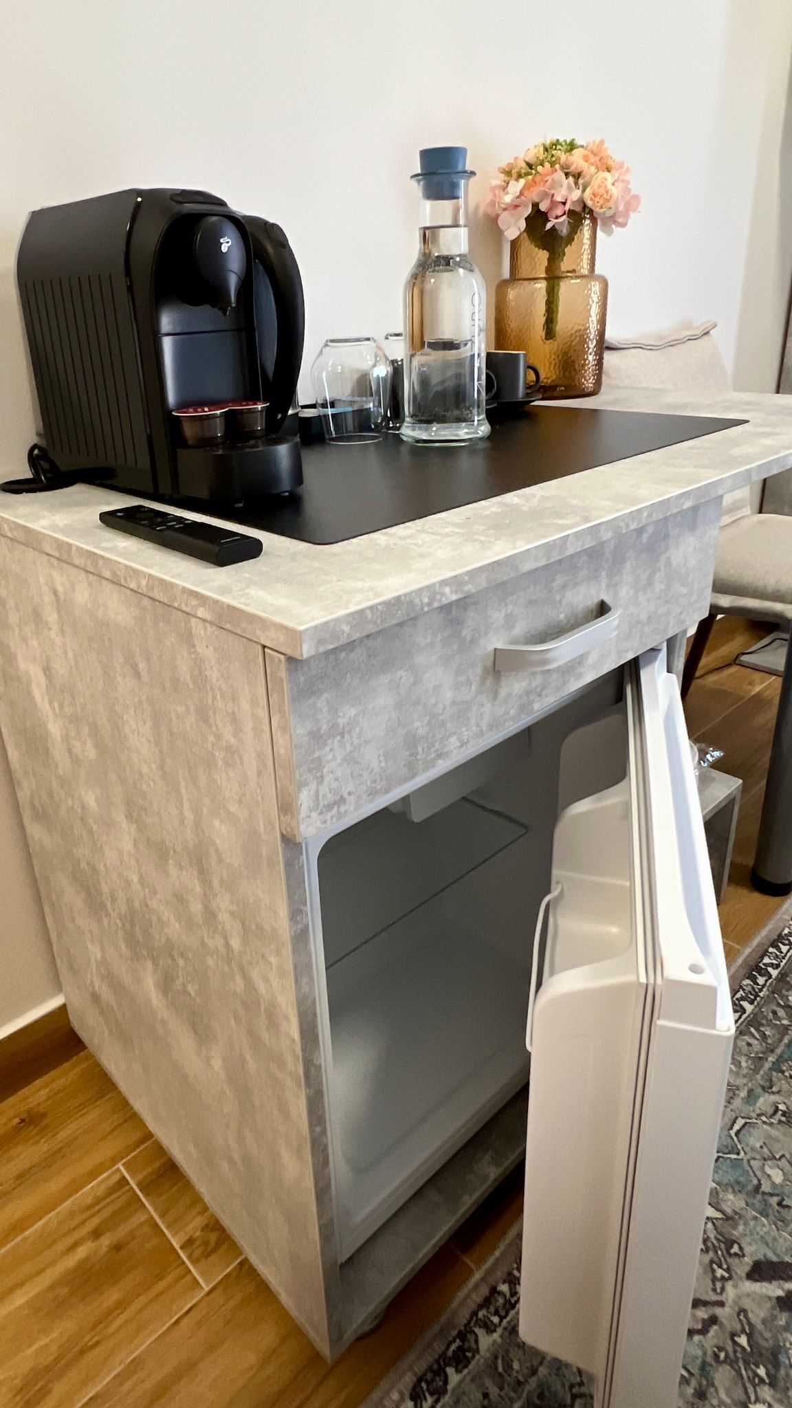 A compact kitchenette with a coffee maker, a bottle, and a vase with flowers.