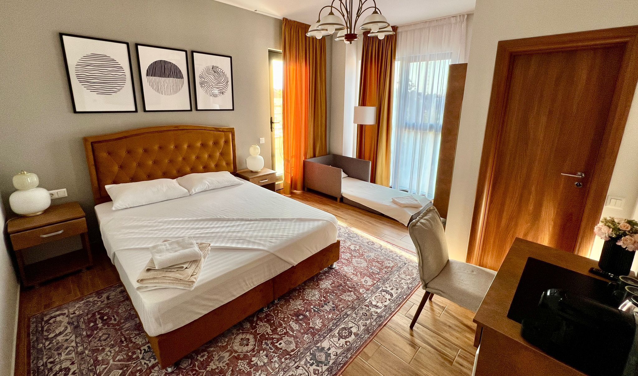 A hotel room with a large bed, a desk with a chair, a small sofa, and a TV. There are framed pictures on the wall, a chandelier, and a window with curtains.