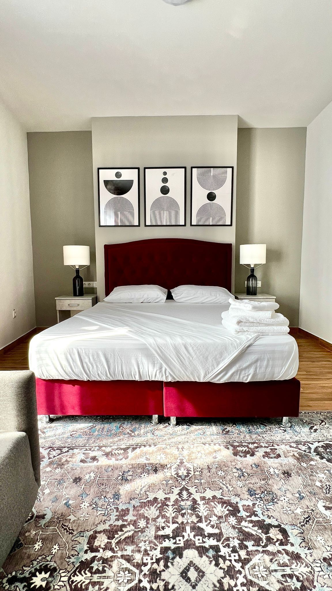 Habitación de hotel con una cama grande, dos mesitas de noche con lámparas, cabecera roja y tres obras de arte enmarcadas en la pared.