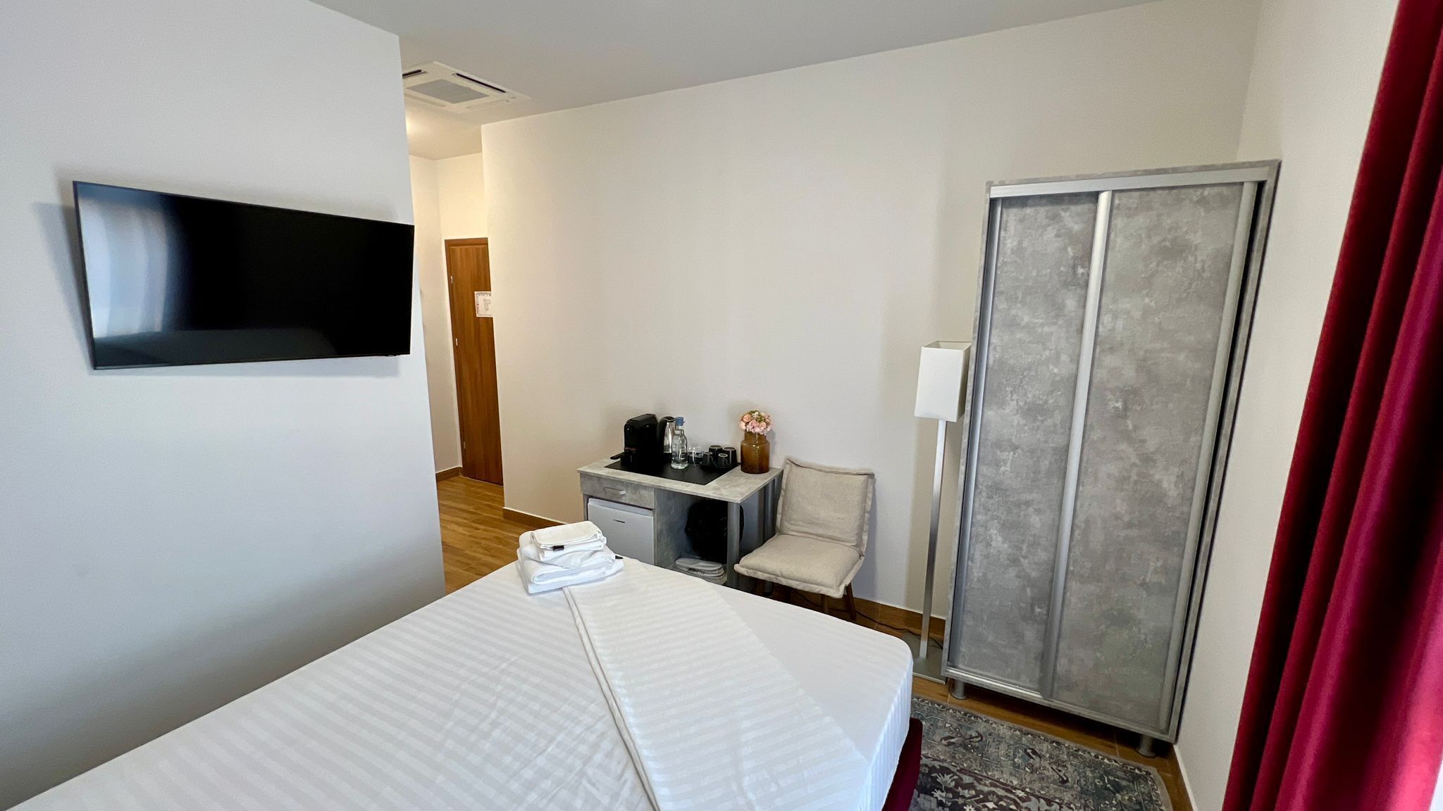 A hotel room with a bed, a desk with a chair, a television, a wardrobe, and a red curtain.