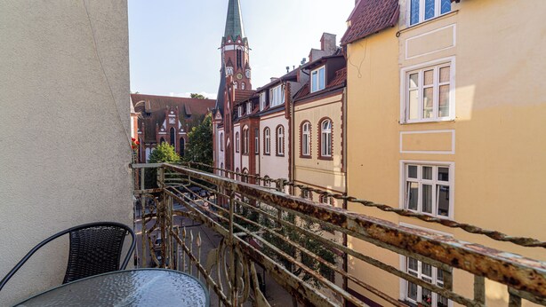 A balcony with a metal railing overlooking a street with historic buildings and a church spire. - Red by Grand Apartments (Sopot)