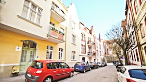A narrow cobblestone street lined with multi-story buildings, parked cars, and a green door on the left. - Red by Grand Apartments (Sopot)
