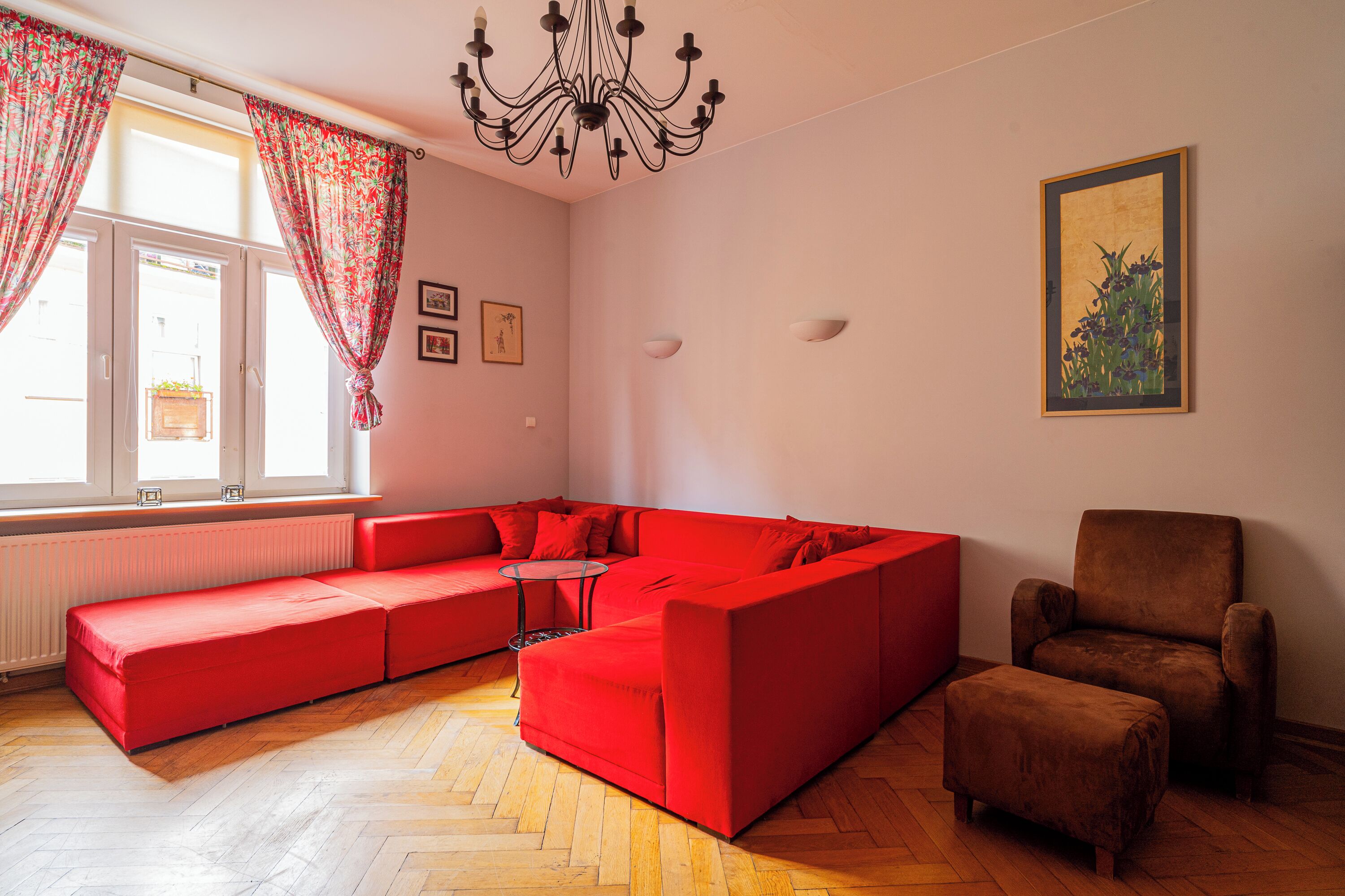 A living room with a red sectional sofa, a brown armchair, and a small ottoman. There is a chandelier, a window with curtains, and a framed picture on the wall.