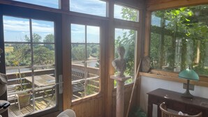 Ein Zimmer mit großen Fenstern, die auf einen Garten und einen Fluss blicken. Es steht eine Büste auf einem Sockel, ein Holztisch mit einer Lampe, und verschiedene Gegenstände sind verstreut.