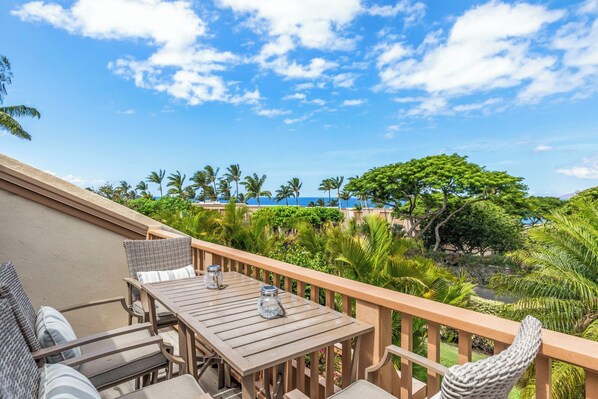 A wooden deck with a table and chairs, overlooking a tropical landscape with palm trees and a view of the ocean.