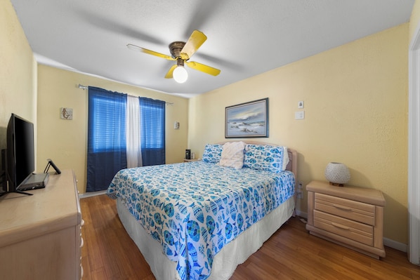Un dormitorio con cama, ventilador de techo, mesita de noche, cómoda, televisor y ventana con cortinas.