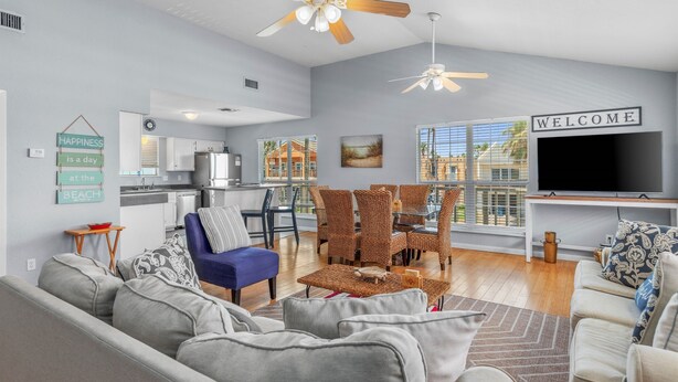 A modern living room with a sofa, coffee table, and a TV. There is a dining area with wicker chairs and a kitchen in the background. - 121 E Aries - Angel's Paradise (South Padre Island)