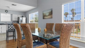 A dining area with wicker chairs and a glass table, kitchen cabinets, and a painting on the wall. - 121 E Aries - Angel's Paradise (South Padre Island)