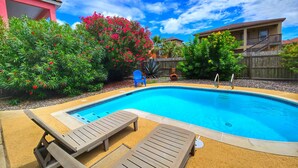 A swimming pool with lounge chairs, surrounded by lush greenery and a wooden fence. - 121 E Aries - Angel's Paradise (South Padre Island)
