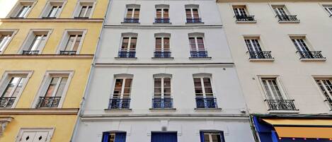 Un immeuble à plusieurs étages doté d’une porte bleue au centre, de façades blanches et jaunes, et de balcons avec des garde-corps noirs.