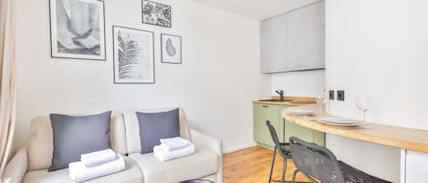 Un salon moderne comprenant un canapé, deux fauteuils, une petite table et un coin cuisine avec un comptoir et des tabourets.