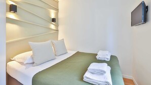 Un lit bien fait, avec du linge de lit blanc et un plaid vert, un téléviseur fixé au mur et une pile de serviettes pliées sur une table de chevet.