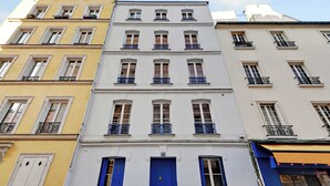 Un immeuble à plusieurs étages doté d’une porte bleue au centre, de façades blanches et jaunes, et de balcons avec des garde-corps noirs.