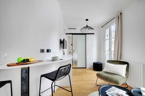 A modern kitchen with a white countertop, two black bar stools, a wooden cutting board with fruits, and a hanging light fixture. - Cosy Studio - 4P/1B  - Gare de Lyon / Bastille (Paris)