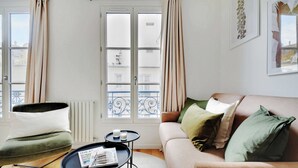 Un salón con un sofá, una mesita redonda con una revista, y una silla. Hay una ventana con cortinas, un radiador y un balcón con barandilla.