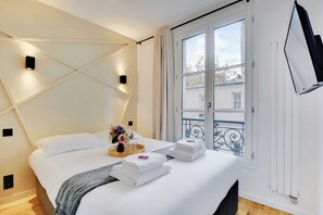 A neatly made bed with white linens, a bedside table with a vase of flowers, and a window with a view of the outside.