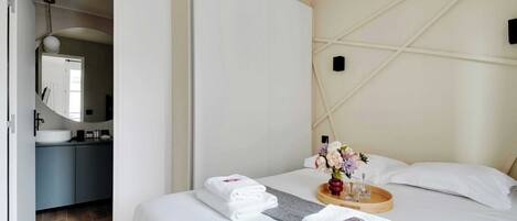 Une chambre d’hôtel avec un lit, une table de chevet sur laquelle se trouve un plateau avec des articles de toilette, un miroir et une salle de bain visible à travers une porte ouverte.