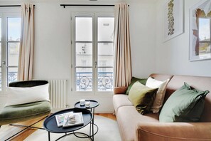 A living room with a sofa, a small round table with a magazine, and a chair. There is a window with curtains, a radiator, and a balcony with a railing. - Cosy Studio - 4P/1B  - Gare de Lyon / Bastille (Paris)