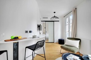 A modern kitchen with a white countertop, two black bar stools, a wooden cutting board with fruits, and a hanging light fixture. - Cosy Studio - 4P/1B  - Gare de Lyon / Bastille (Paris)