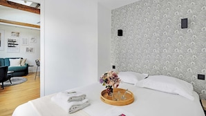 Una habitación de hotel moderna con una cama grande, una bandeja de madera con toallas y un jarrón de flores, un papel tapiz decorativo y vistas al área de estar contigua.