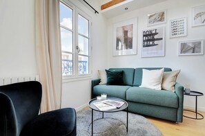 Een moderne woonkamer met een blauwgroene bank, een zwarte stoel, een klein rond tafeltje met een tijdschrift, en een raam met gordijnen.