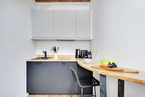 Een compacte keuken met een grijze kast, een houten aanrecht, een zwarte stoel en een fruitschaal met appels en een groene paprika.