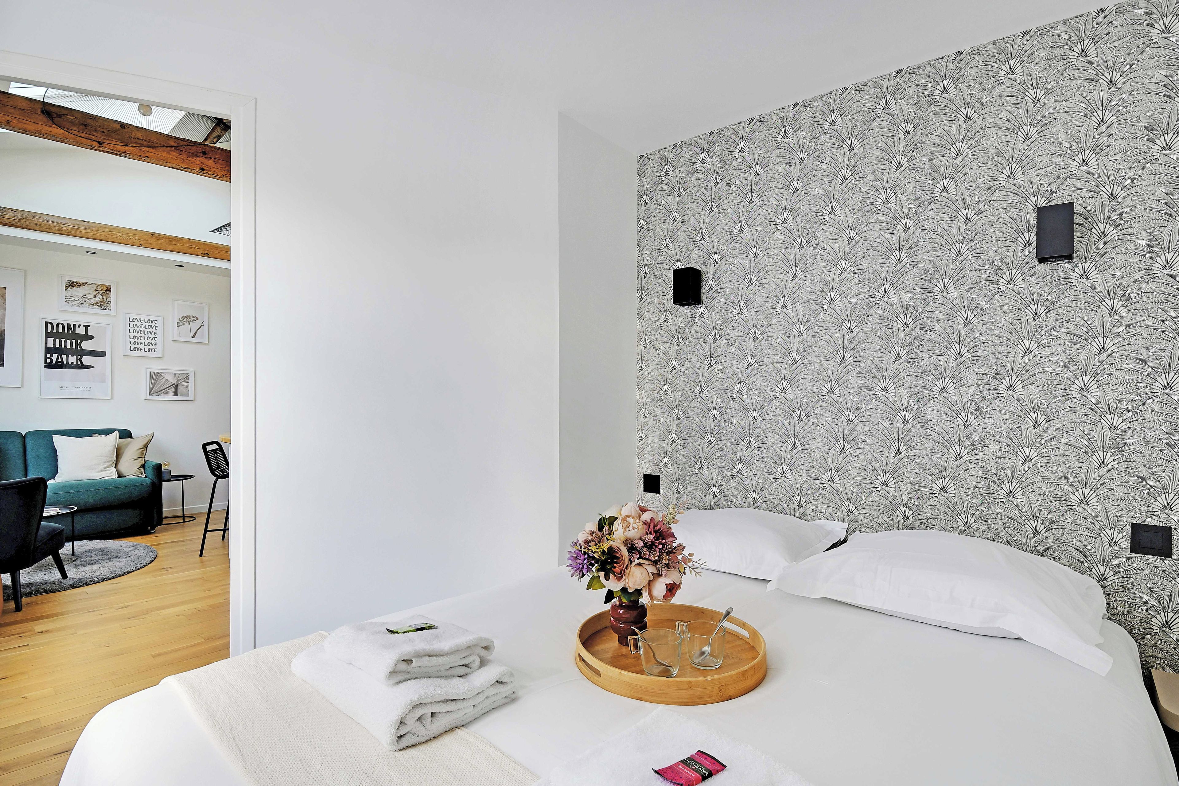 Une chambre d’hôtel moderne avec un grand lit, un plateau en bois contenant des serviettes et un vase de fleurs, un mur décoré d’un motif et une vue sur l’espace de vie adjacent.