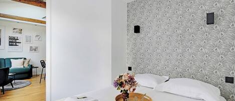 Une chambre d’hôtel moderne avec un grand lit, un plateau en bois contenant des serviettes et un vase de fleurs, un mur décoré d’un motif et une vue sur l’espace de vie adjacent.