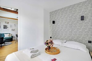 Camera d'albergo moderna con un letto grande, un vassoio di legno con asciugamani e un vaso di fiori, una parete decorata con motivi e vista sull'area soggiorno adiacente.