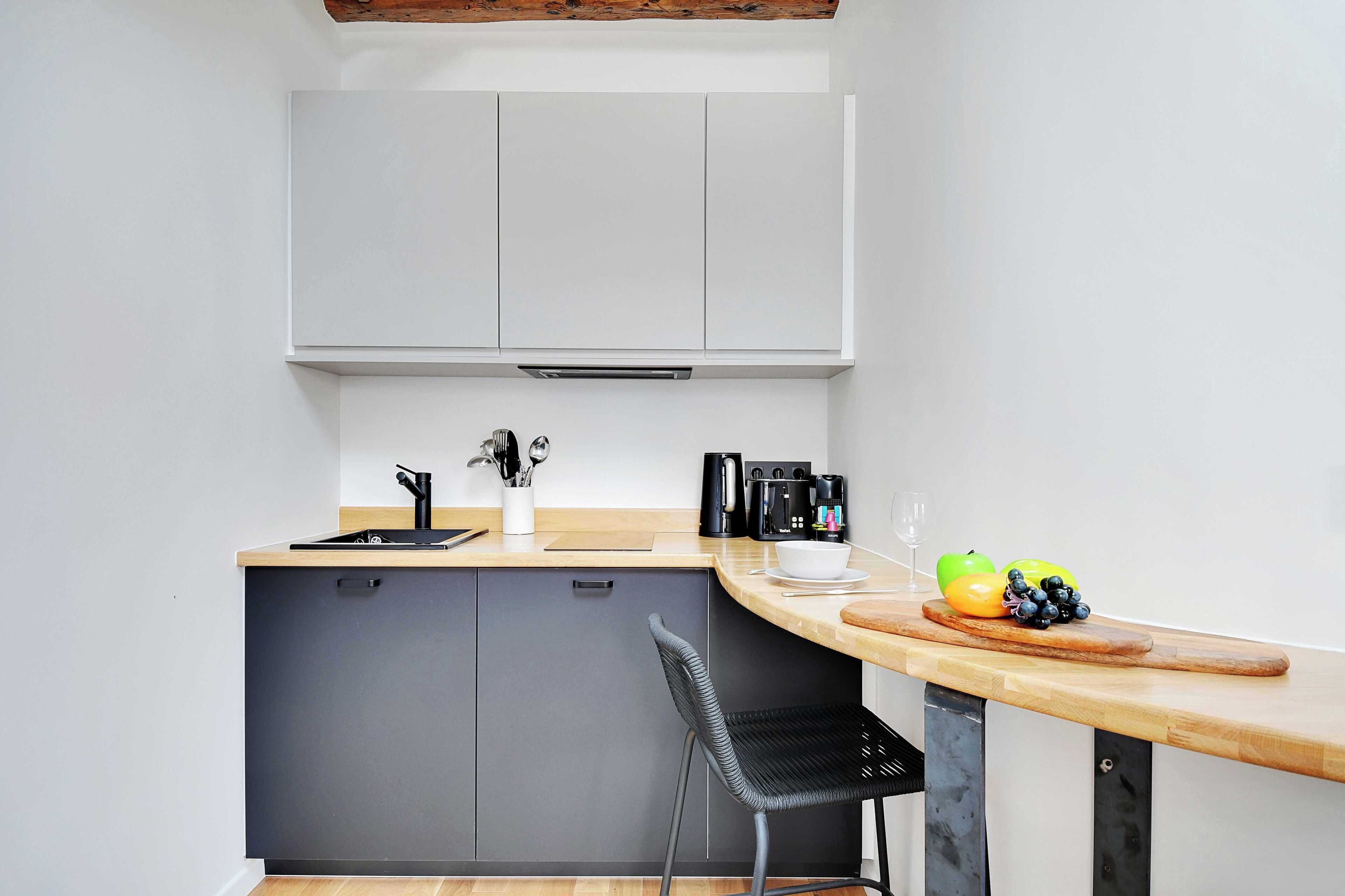 Een compacte keuken met een grijze kast, een houten aanrecht, een zwarte stoel en een fruitschaal met appels en een groene paprika.