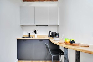 Een compacte keuken met een grijze kast, een houten aanrecht, een zwarte stoel en een fruitschaal met appels en een groene paprika.