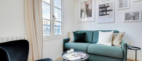 Un salon moderne avec un canapé de couleur turquoise, un fauteuil noir, une petite table ronde sur laquelle repose un magazine, et une fenêtre avec des rideaux.