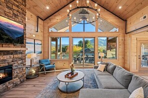 A spacious living room with a stone fireplace, a large sofa, a round coffee table, and a chandelier. - Luxe 6BR w/Swim Spa, Theater, Firepit, Games! (Sevierville)