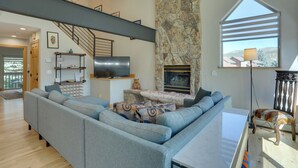 Ein modernes Wohnzimmer mit Kamin aus Stein, einem großen grauen Ecksofa, einem Flachbildfernseher und einer Treppe, die in eine obere Ebene führt.