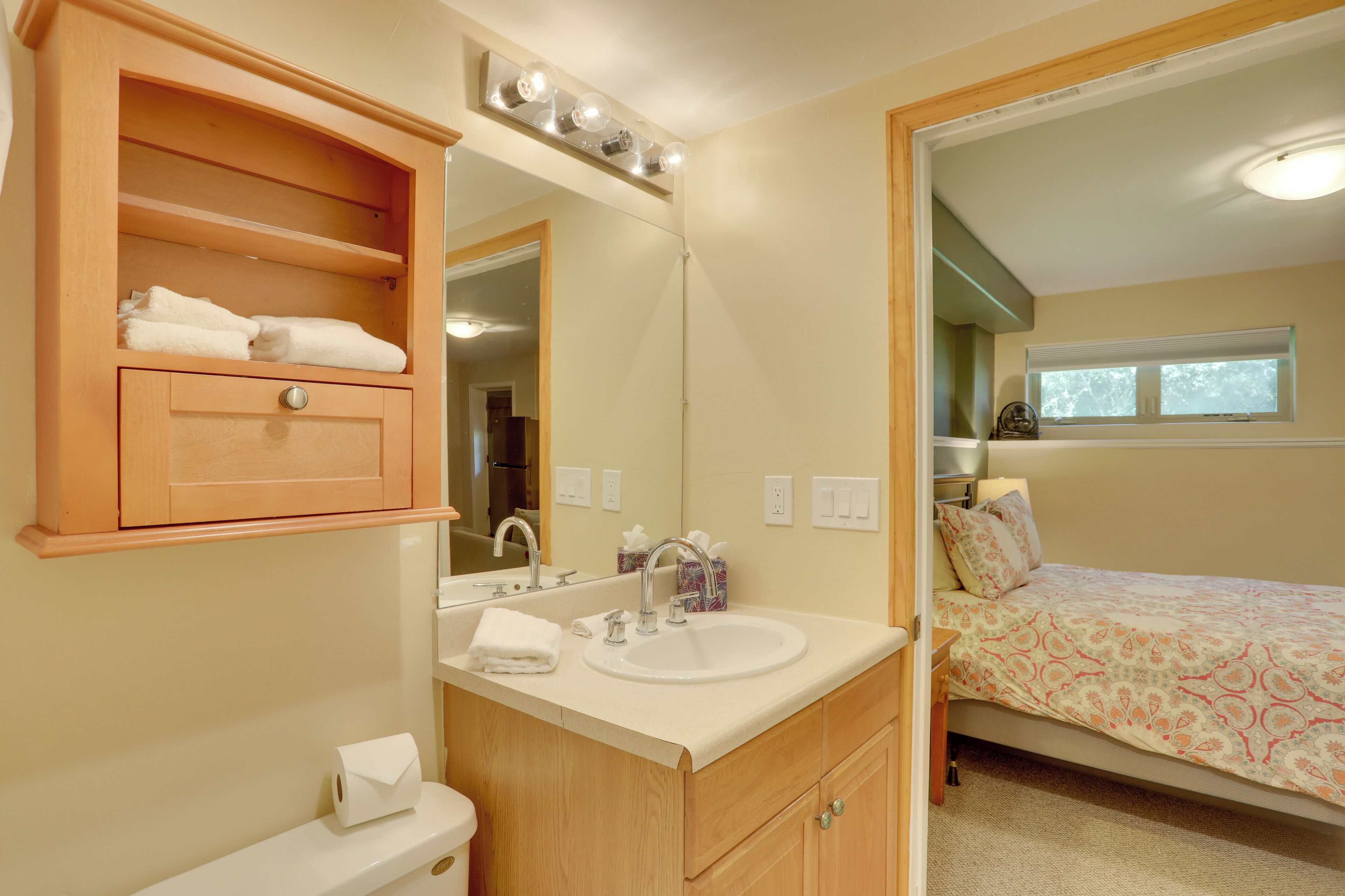 Une salle de bain avec un meuble-lavabo en bois, un évier et un miroir. La pièce adjacente contient un lit recouvert d’une couvre-lit à motifs.