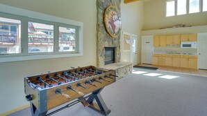 Amplia habitación con mesa de fútbol, chimenea de piedra, zona de cocina y ventanas grandes con vistas al exterior.