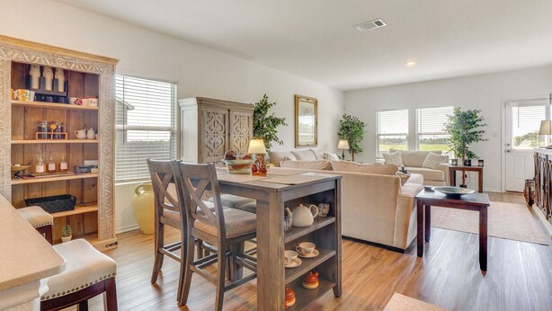 Un salón espacioso con zona de comedor, un conjunto de sofás, un mueble de madera con utensilios de cocina y ventanas amplias que permiten la entrada de luz natural.