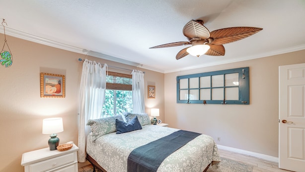 Une chambre à coucher comprenant un lit, des tables de chevet, un ventilateur de plafond, une fenêtre avec des rideaux et une porte.