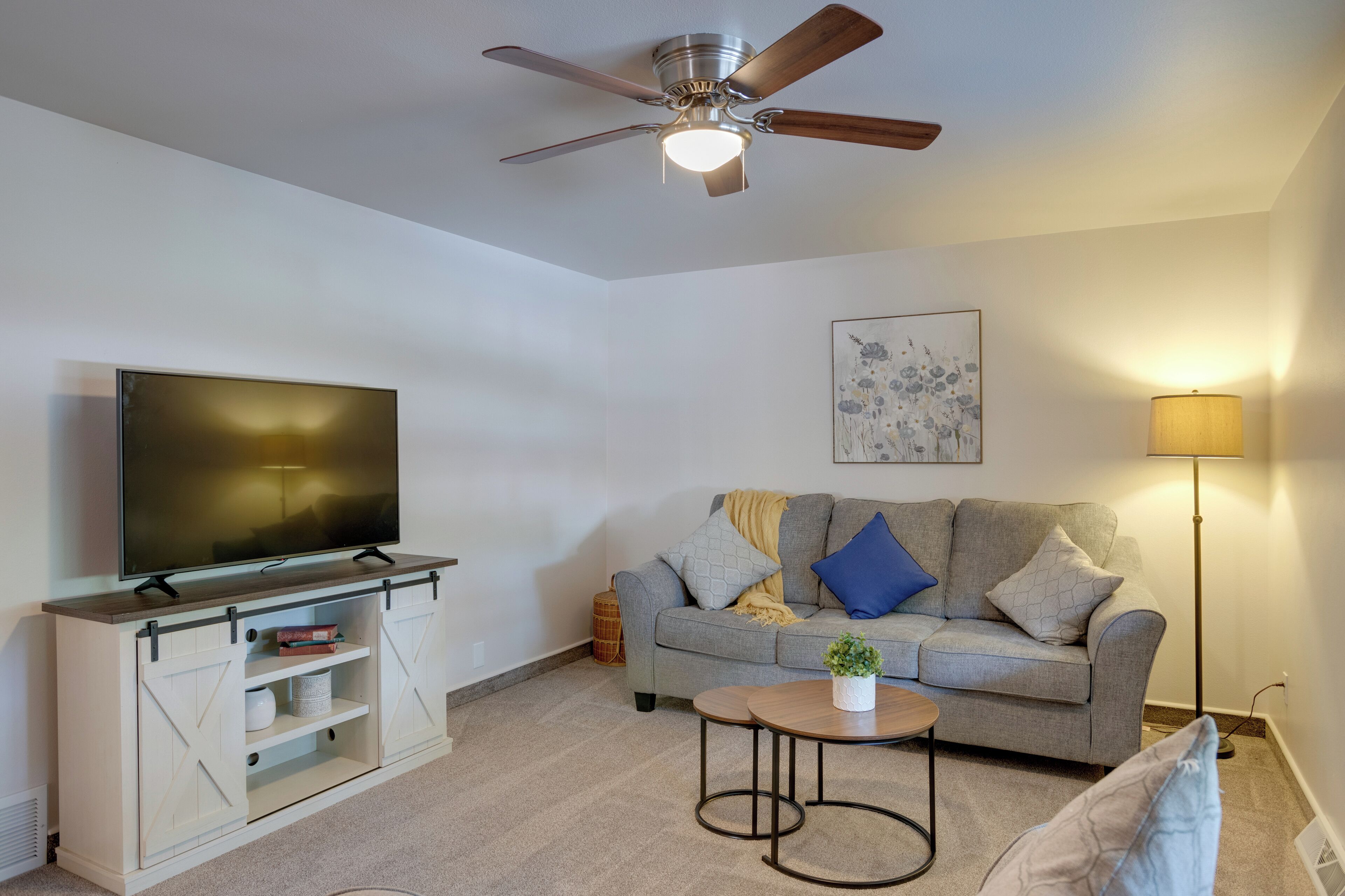 A living room with a flat-screen TV on a wooden cabinet, a grey sofa with cushions, a small round table with a plant, a standing floor lamp, and a ceiling fan.