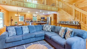 A spacious living room with a large sectional sofa, a wooden staircase, and a kitchen area in the background. - Deck & Private Hot Tub: Cabin in Ellijay! (Ellijay)