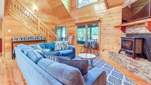 A log cabin interior with a wooden staircase, a blue sofa set, a stone fireplace, and a sign that reads "EAT & DRINK". - Deck & Private Hot Tub: Cabin in Ellijay! (Ellijay)