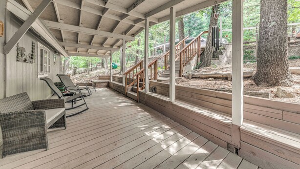 Een overdekt houten terras met lounge fauteuils, een trap naar een terras en uitzicht op het omliggende bos.