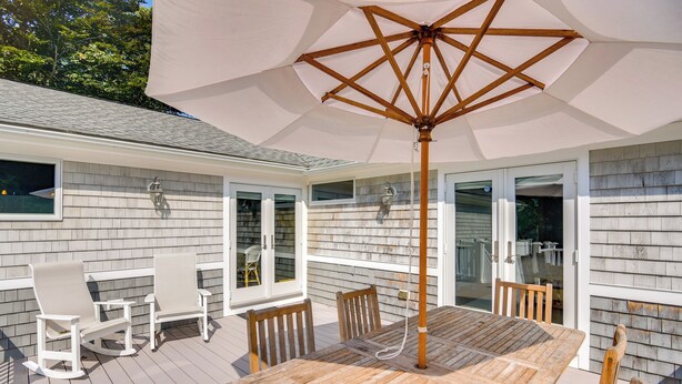 A wooden deck with a table and chairs, a large umbrella, and a house with white siding and windows. - Coastal-modern Home < 3 Mi to Charlestown Beach! (South Kingstown)
