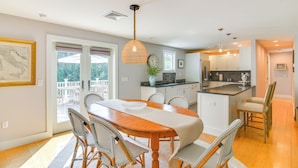 A spacious dining area with a wooden table and wicker chairs, a kitchen with white cabinets, and a view through the glass door. - Coastal-modern Home < 3 Mi to Charlestown Beach! (South Kingstown)
