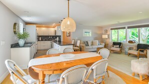 A spacious living area with a dining table, wicker chairs, and a large window providing a view of greenery. - Coastal-modern Home < 3 Mi to Charlestown Beach! (South Kingstown)