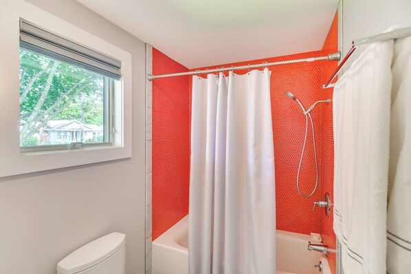 A bathroom with a white bathtub, a shower with a red tiled wall, and a window with a view of trees and houses. - Coastal-modern Home < 3 Mi to Charlestown Beach! (South Kingstown)